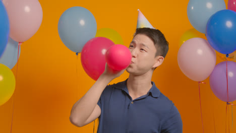Studioporträt-Eines-Mannes-Mit-Partyhut,-Der-Seinen-Geburtstag-Feiert-Und-Einen-Ballon-In-Die-Luft-Sprengt